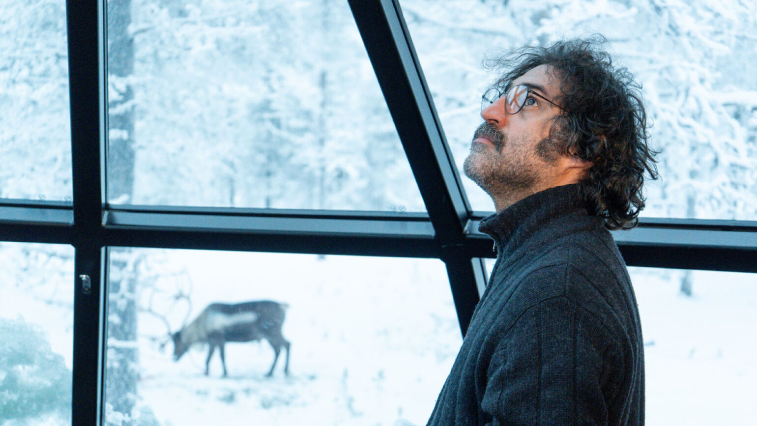 雪景色が広がる窓の外を、眼鏡をかけた男性が室内から見上げている画像。