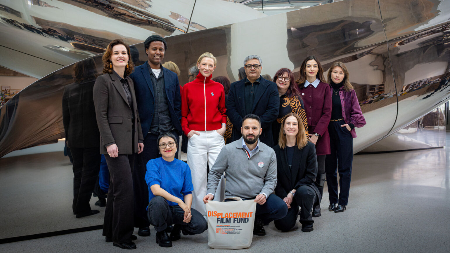 ケイト・ブランシェットさんを中心に、「DISPLACEMENT FILM FUND」と書かれたバッグを持つ関係者たちが銀色のオブジェの前で撮影した集合写真の画像。