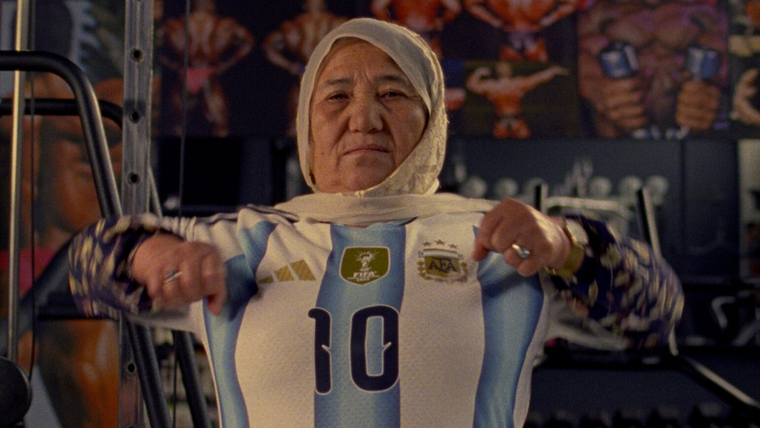 サッカーアルゼンチン代表のユニフォームを着て、ジムのトレーニング機器を前に力強い表情を浮かべる年配女性の画像。