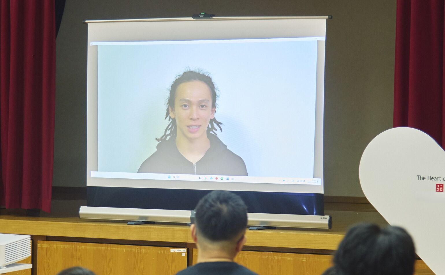 平野歩夢選手がビデオメッセージを通じて挨拶している様子が、プロジェクターのスクリーンに映し出されている画像