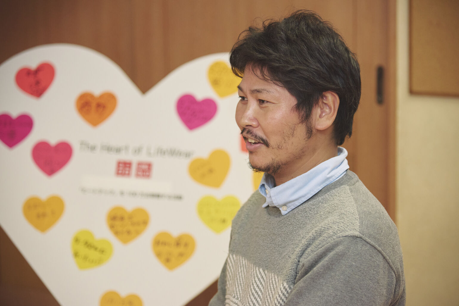 メッセージが貼られたハート型のボードを背景に、男性が左側を向いて話している様子の画像
