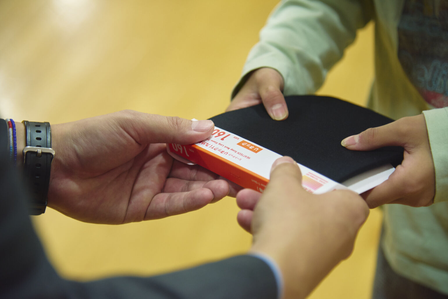 薄緑色のシャツを着た子供が、両手で黒いヒートテックのパッケージを受け取っている手元のクローズアップ画像。