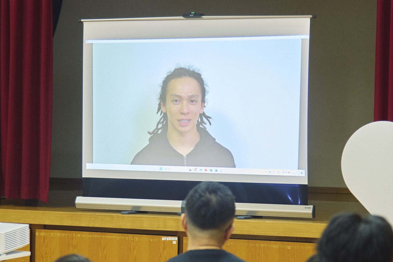 平野歩夢選手がビデオメッセージを通じて挨拶している様子が、プロジェクターのスクリーンに映し出されている画像