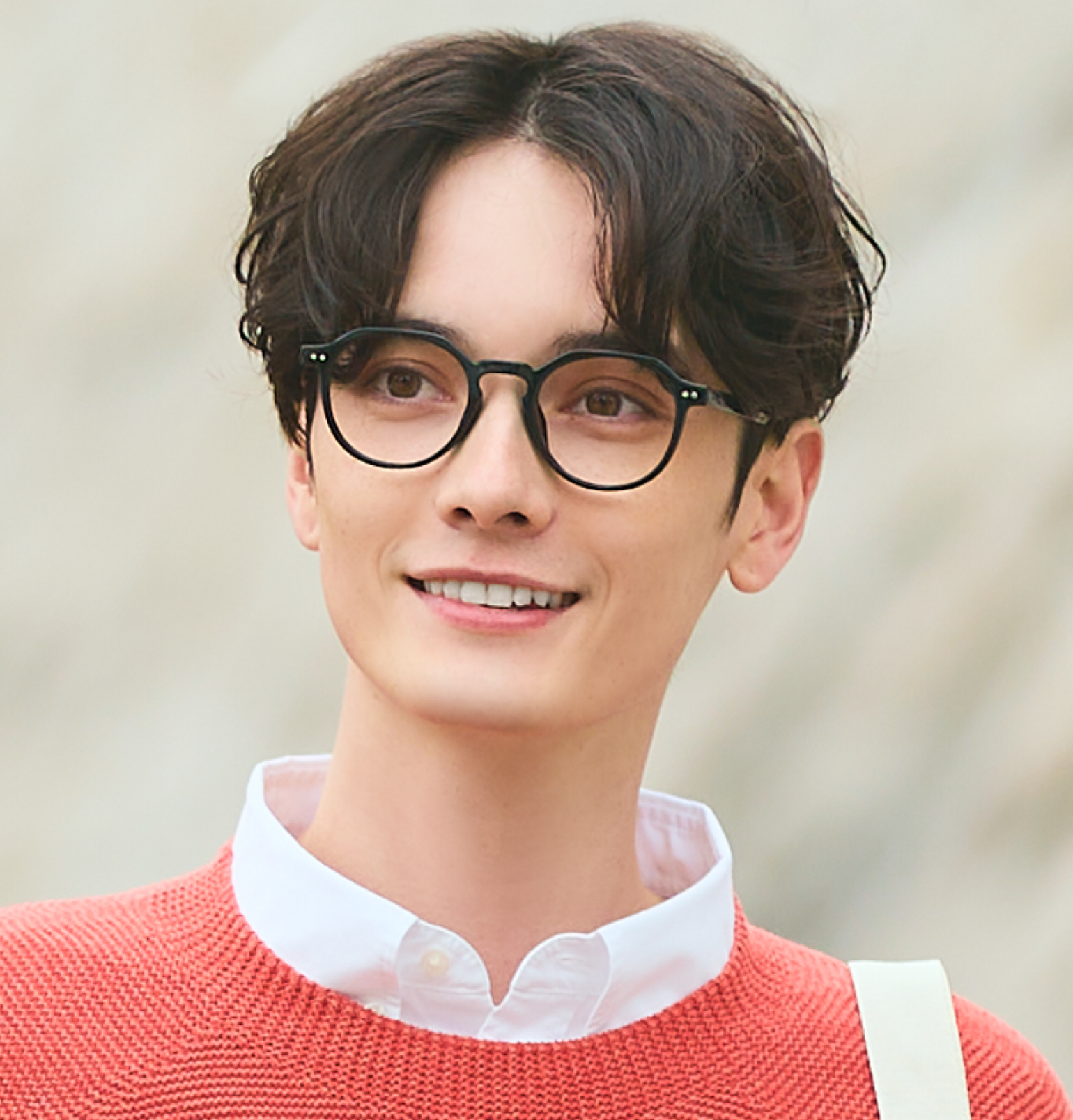 多角形フレームのクラシックな眼鏡をかけた男性が、オレンジ色のニットを着用し、明るい屋外で斜め前方を見つめている画像。